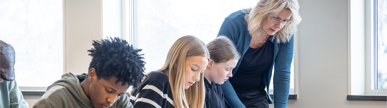 En lärare med ljust hår undervisar tre elever som sitter och arbetar.